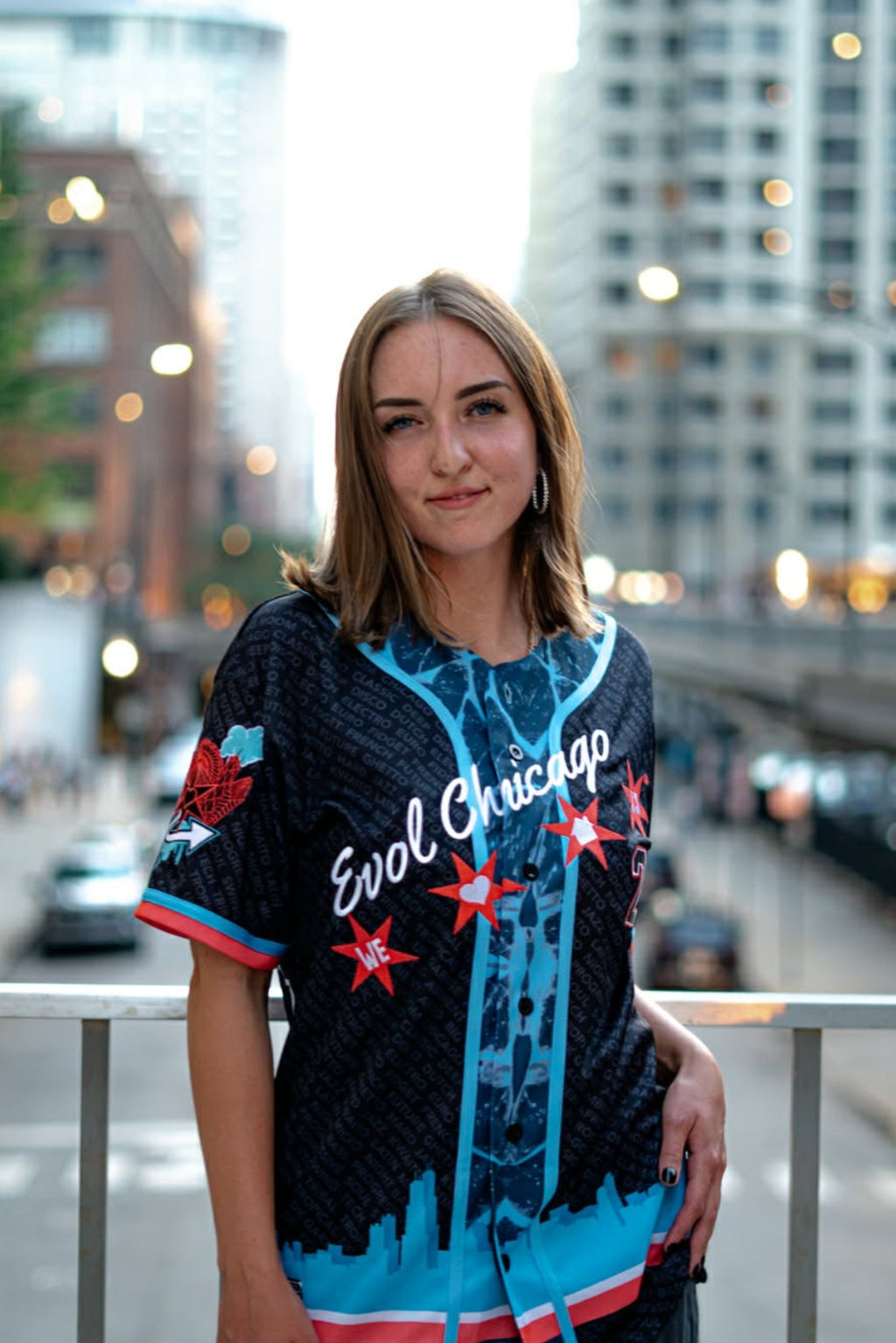 A woman stands on an urban bridge wearing a black short-sleeve baseball-style shirt with blue and red graphics, including the text 'Evol' Chicago' and star motifs, We Love House Music paired with dark cargo pants. The background shows a blurred city street with tall buildings and streetlights.