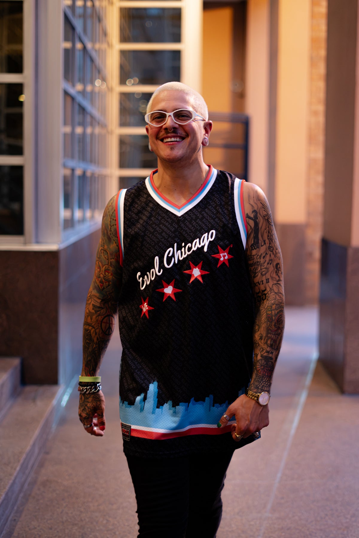A smiling man with a shaved head and tattoos wears a black we love house music Chicago basketball jersey with red and white accents, standing in a hallway.